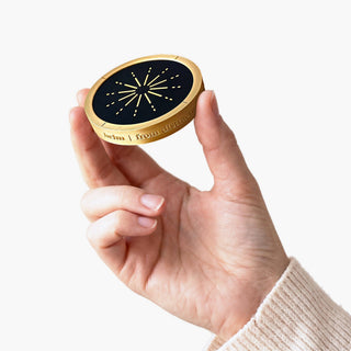 Fingertips hold the brass Helm up so its black face, gold 12-point burst and engraved rim show, on a white background. - BestSelf Co.